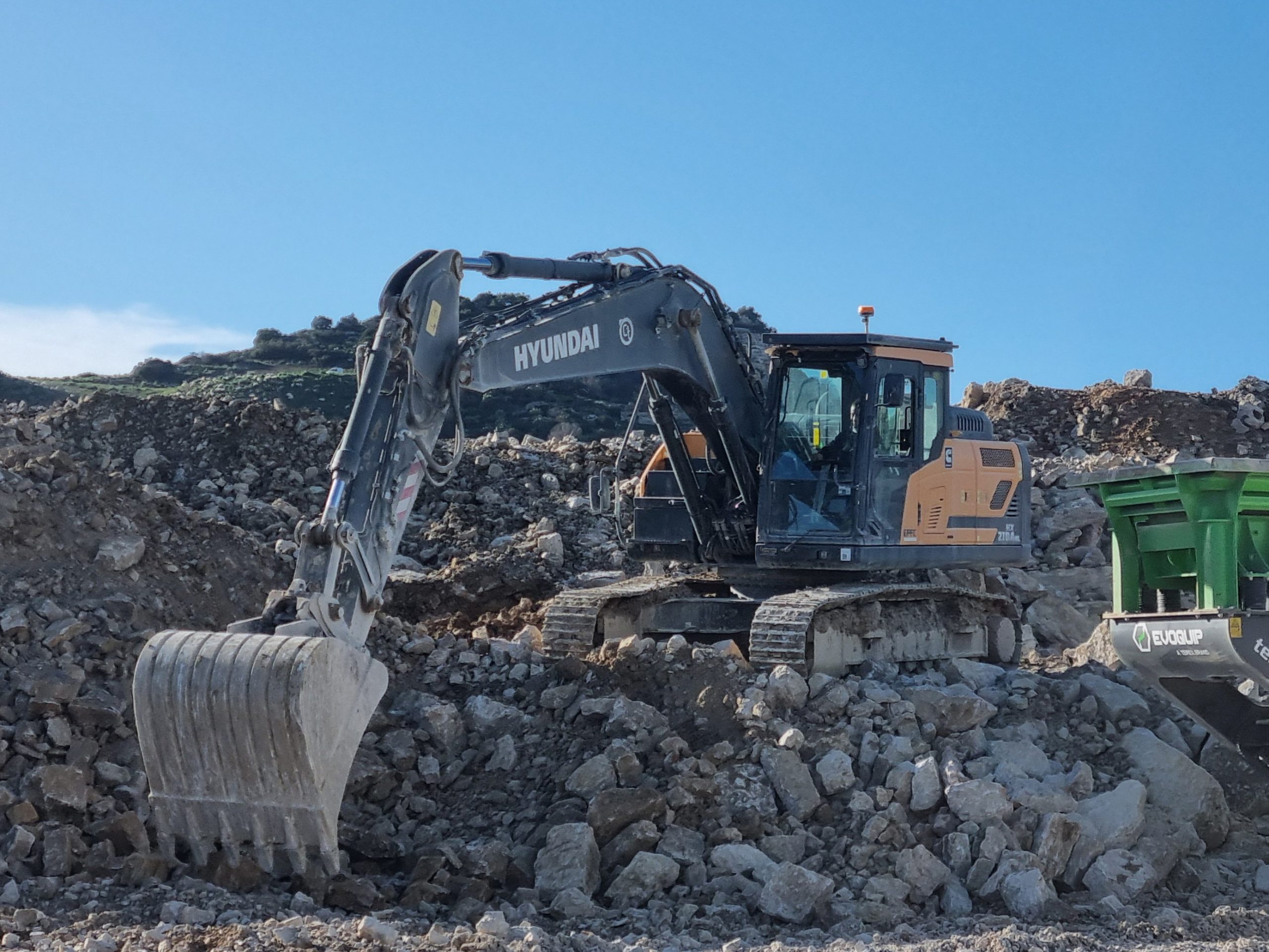 Un escavatore cingolato Hyundai HX210ANL impegnato nella movimentazione di blocchi di roccia e materiale all'interno della cava.