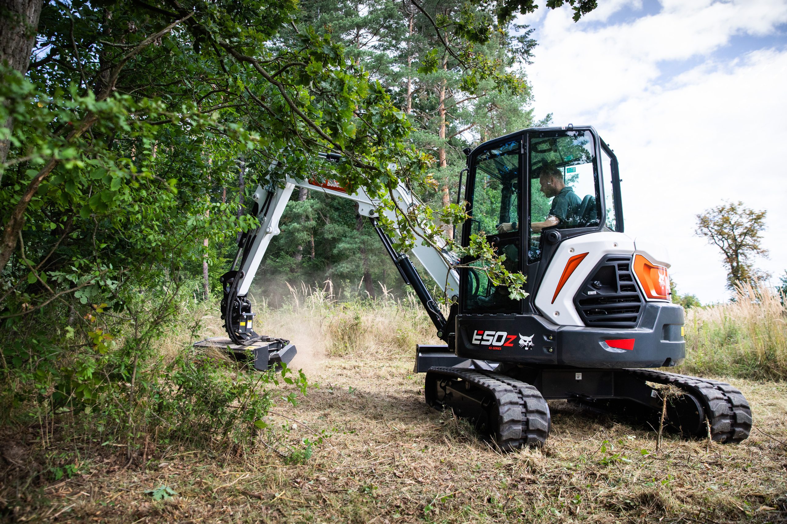 Miniescavatore Bobcat E50z con trinciatutto al lavoro per sfalciare l'erba alta e la vegetazione ai margini del bosco, creando corridoi di sicurezza antincendio.